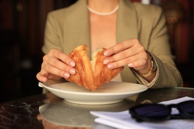 Photo of Woman breaking tasty croissant at table in cafeteria, closeup