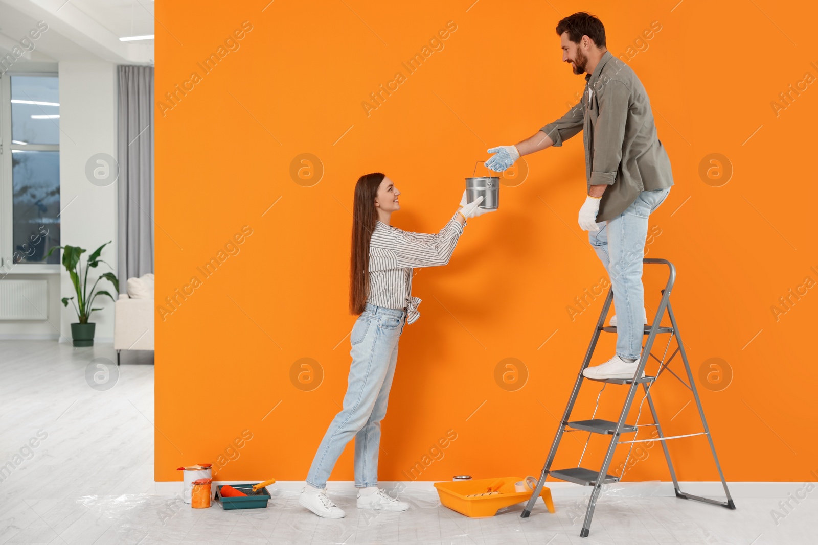 Photo of Woman giving man can of paint near orange wall indoors. Interior design