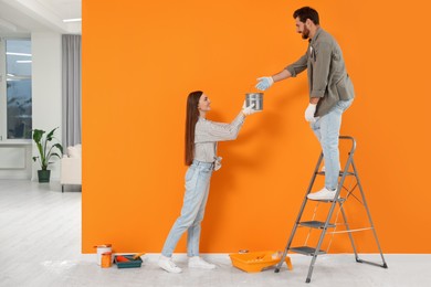 Photo of Woman giving man can of paint near orange wall indoors. Interior design