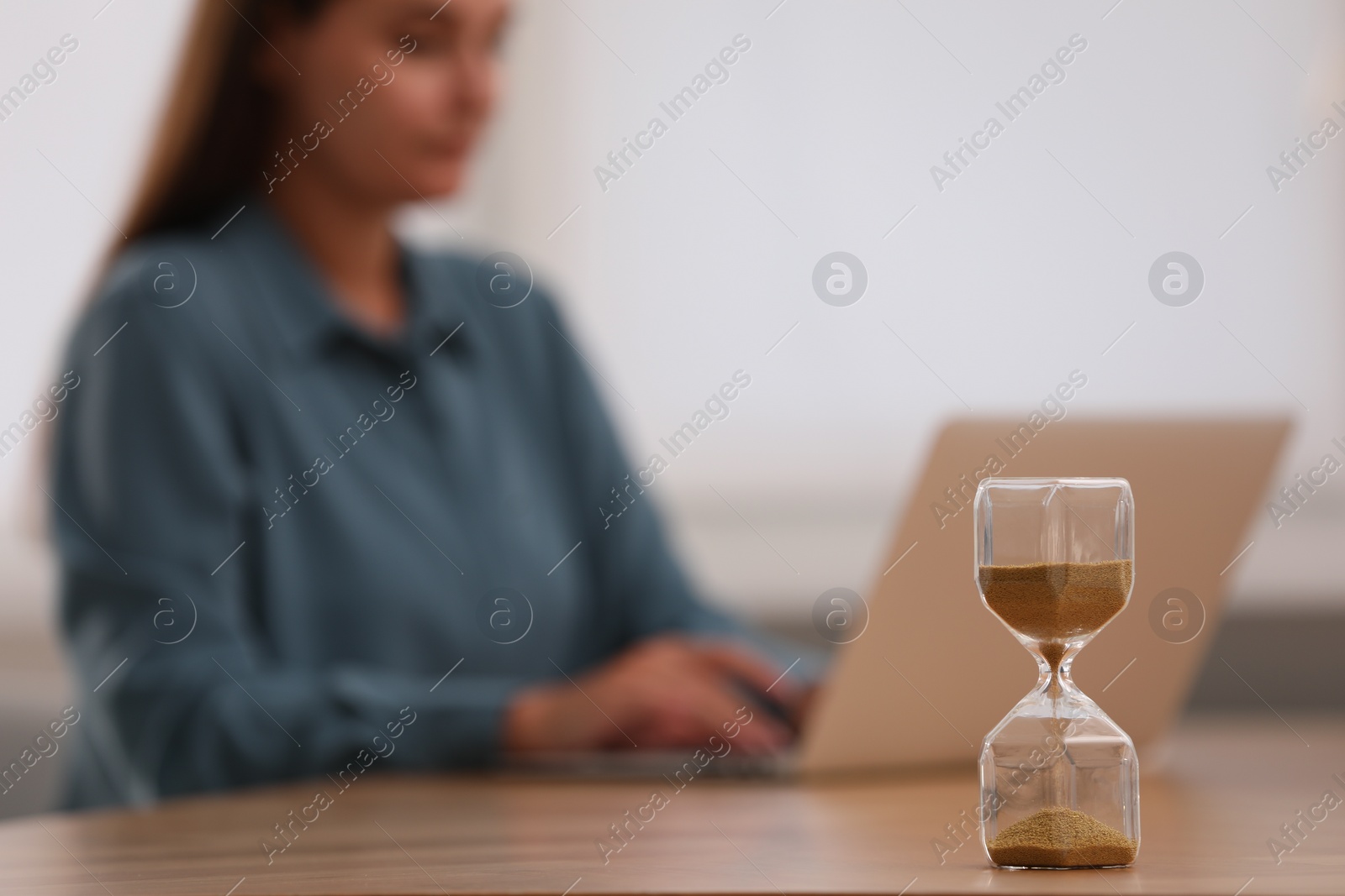 Photo of Hourglass with flowing sand on table. Woman using laptop indoors, selective focus