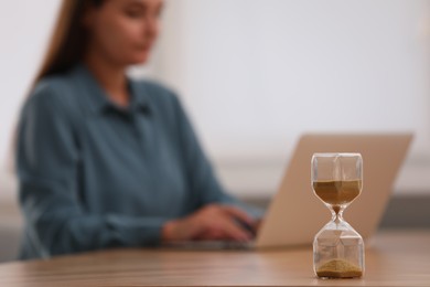 Hourglass with flowing sand on table. Woman using laptop indoors, selective focus