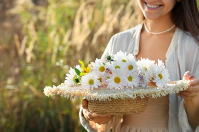 Woman holding straw hat with beautiful wild flowers outdoors, closeup. Space for text