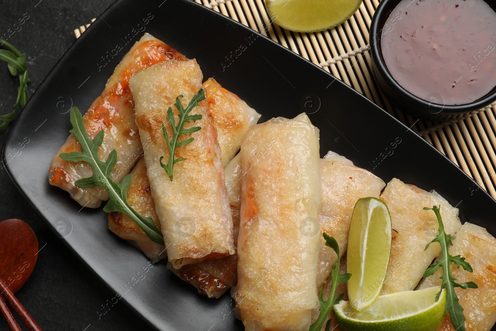 Photo of Tasty fried spring rolls, arugula, lime and sauce on dark textured table, flat lay