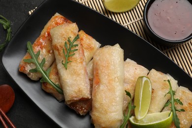 Photo of Tasty fried spring rolls, arugula, lime and sauce on dark textured table, flat lay