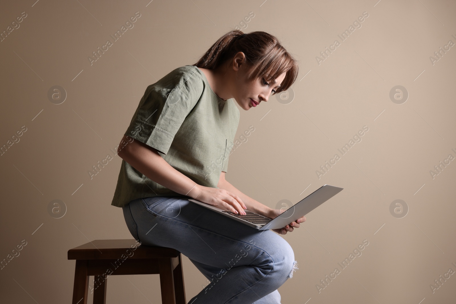 Photo of Young woman with poor posture using laptop while sitting on stool against beige background