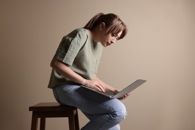 Young woman with poor posture using laptop while sitting on stool against beige background