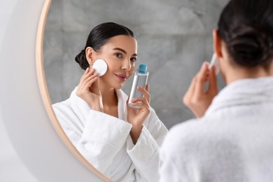 Beautiful woman removing makeup with cotton pad near mirror indoors