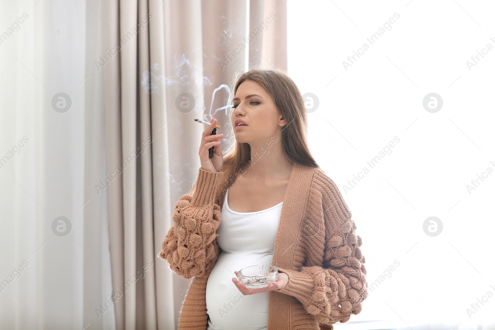 Photo of Young pregnant woman smoking cigarette at home. Harm to unborn baby