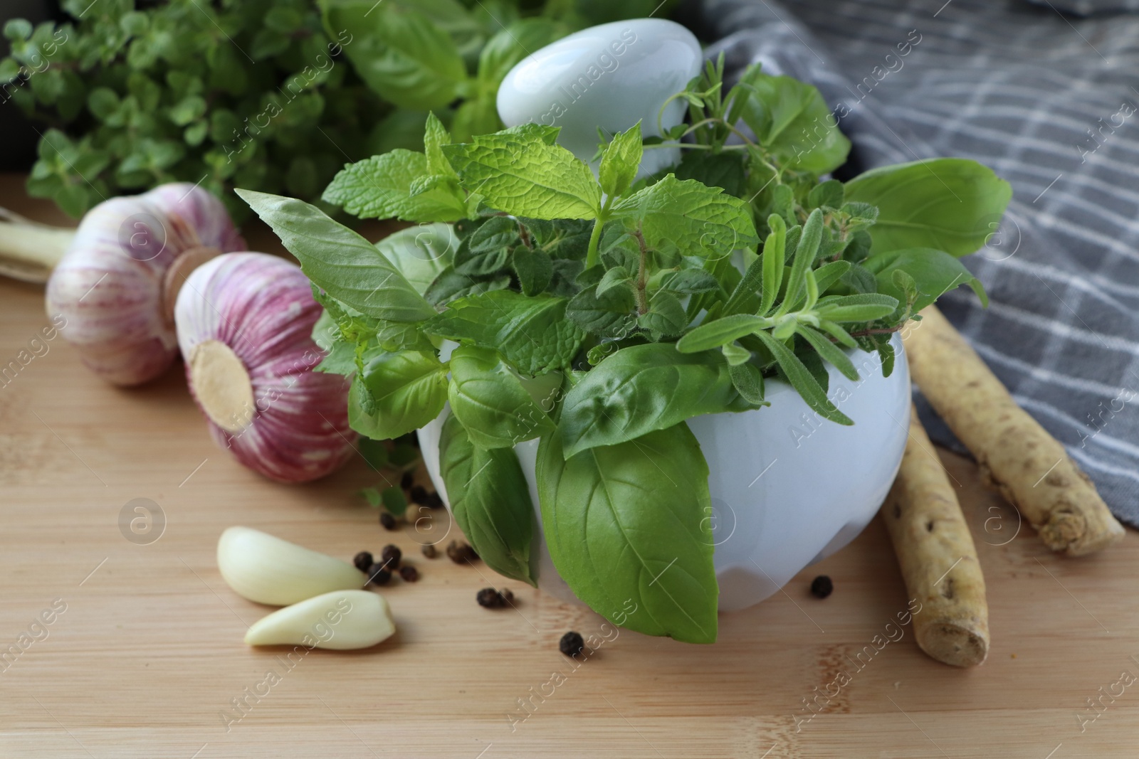 Photo of Mortar with different fresh herbs near garlic, horseradish roots and black peppercorns on wooden table, closeup