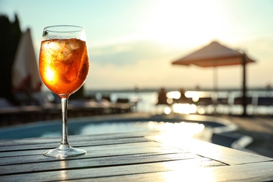Glass of fresh summer cocktail on wooden table near swimming pool outdoors at sunset. Space for text