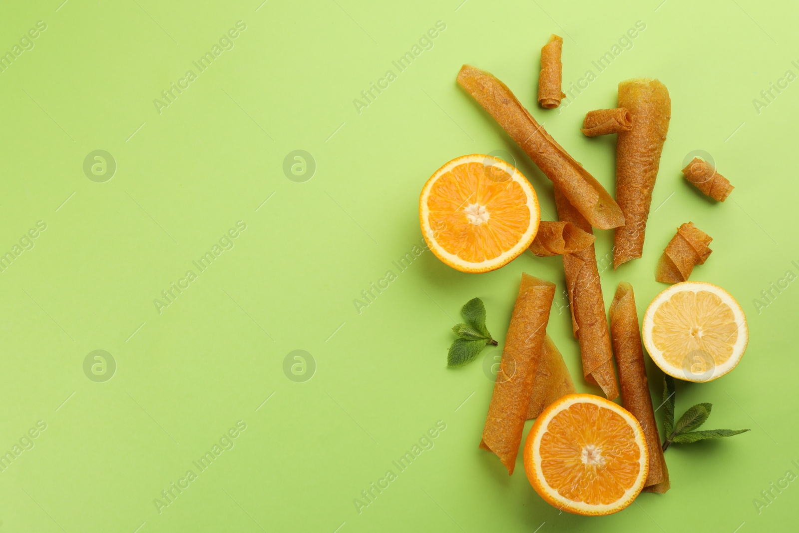 Photo of Delicious fruit leather rolls, mint, lemon and orange on green background, flat lay. Space for text