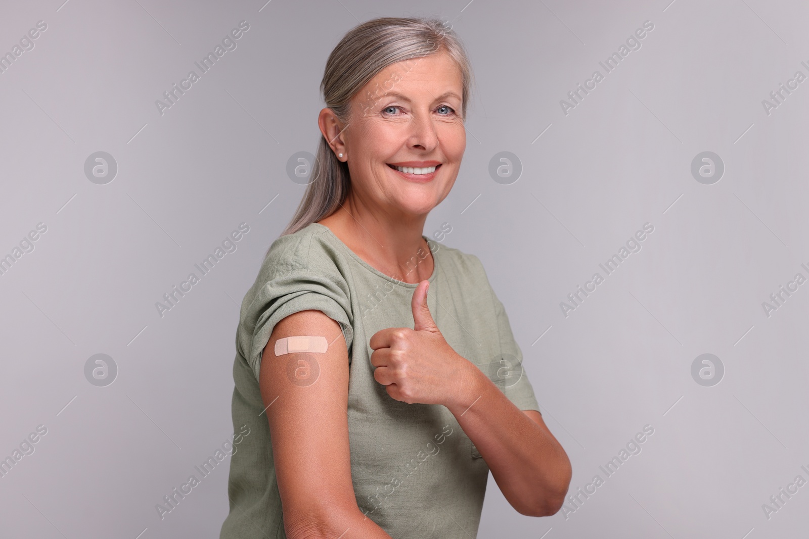 Photo of Senior woman with adhesive bandage on her arm after vaccination showing thumb up against light grey background