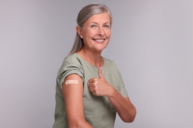 Senior woman with adhesive bandage on her arm after vaccination showing thumb up against light grey background