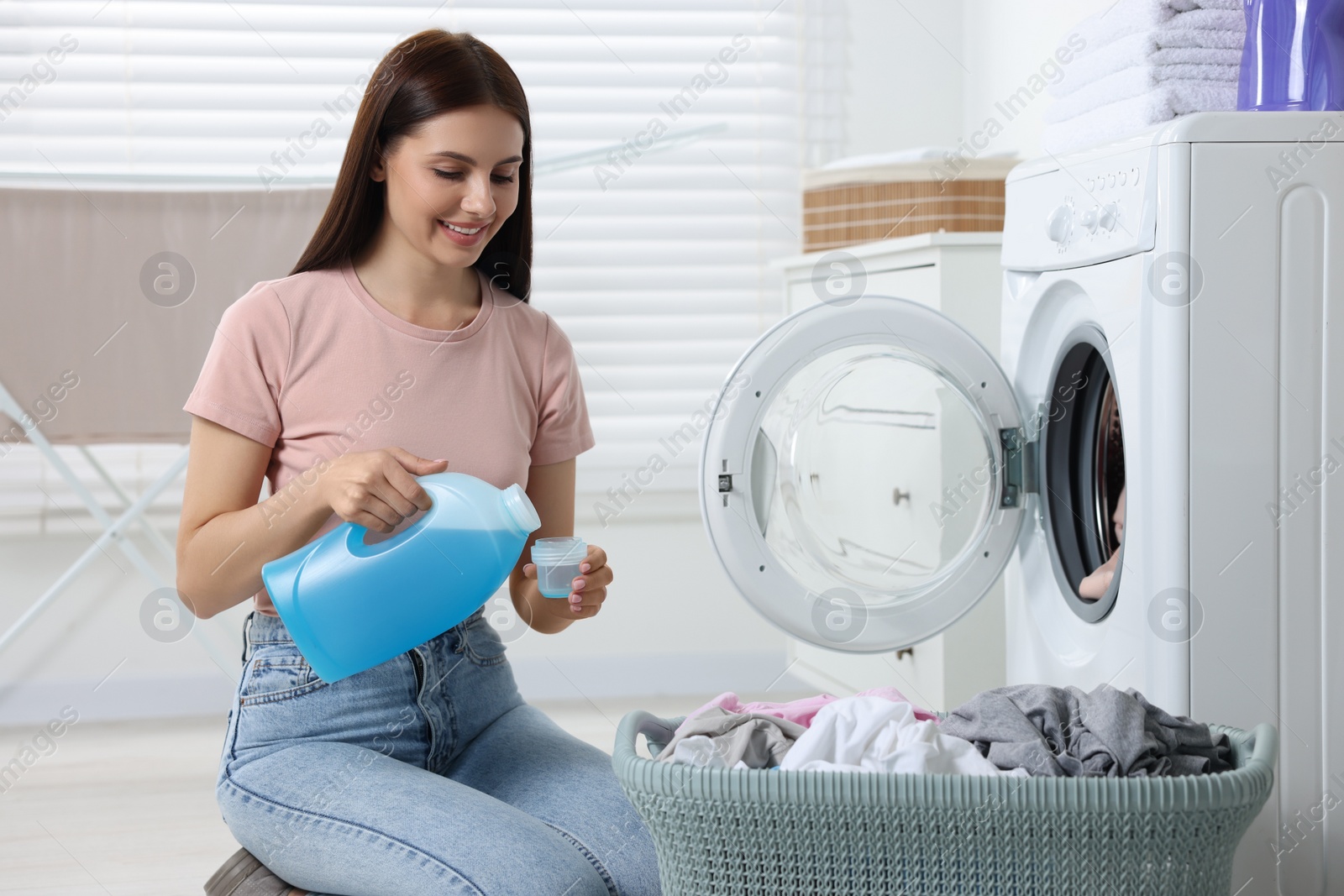 Photo of Woman pouring fabric softener from bottle into cap near washing machine in bathroom