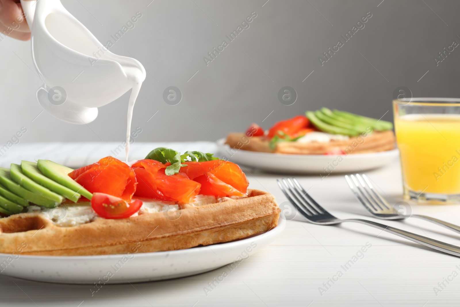 Photo of Woman pouring sauce onto delicious Belgian waffle with salmon, avocado and arugula at white wooden table, closeup. Space for text