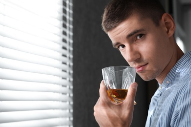 Young man with glass of whiskey near window indoors. Space for text