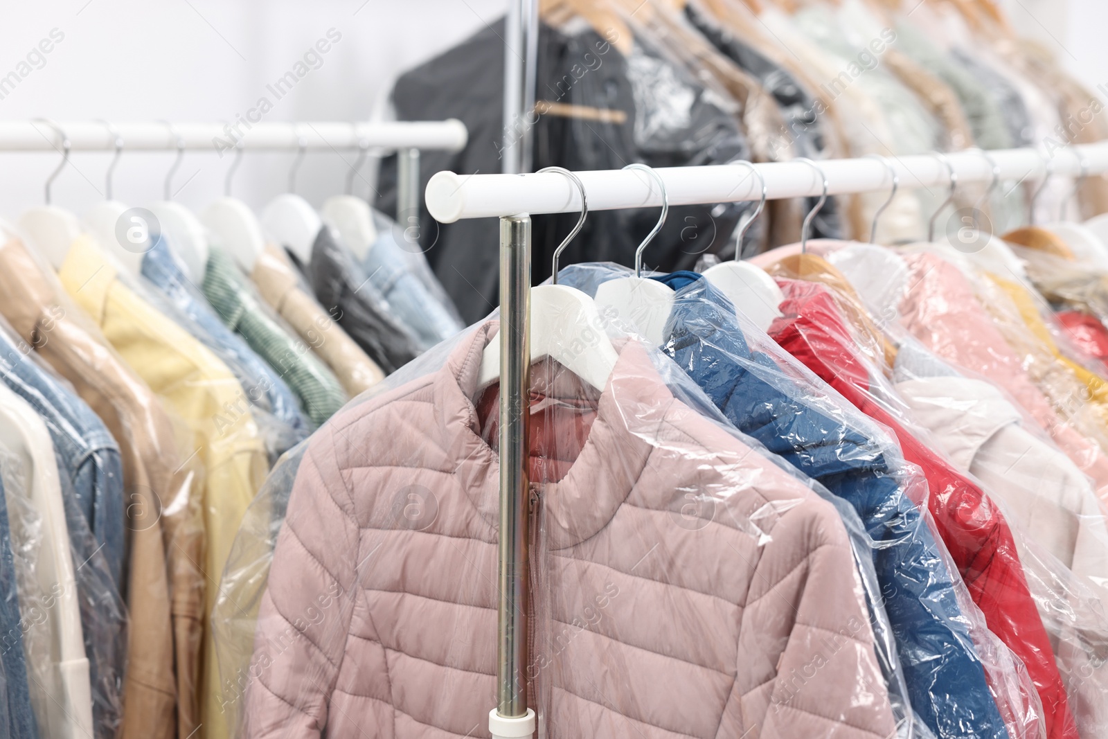 Photo of Dry-cleaning service. Many different clothes in plastic bags hanging on rack indoors, closeup