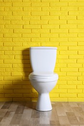 Photo of New clean toilet bowl near yellow brick wall indoors. Interior design