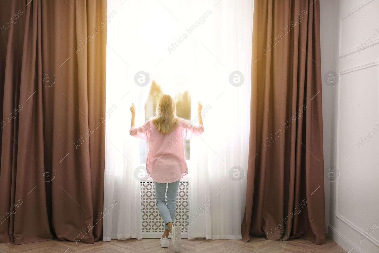 Photo of Young woman opening window curtains at home, back view