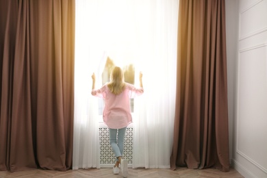 Young woman opening window curtains at home, back view