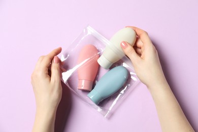 Photo of Cosmetic travel kit. Woman putting small bottle with personal care product into plastic bag against violet background, top view