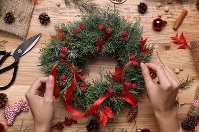 Photo of Florist making beautiful Christmas wreath with berries and red ribbon at wooden table, top view