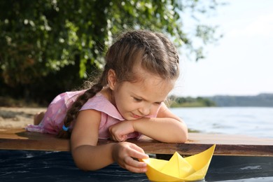 Cute little girl playing with paper boat on wooden pier near river