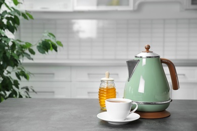 Photo of Modern electric kettle, cup of tea and honey on grey table in kitchen