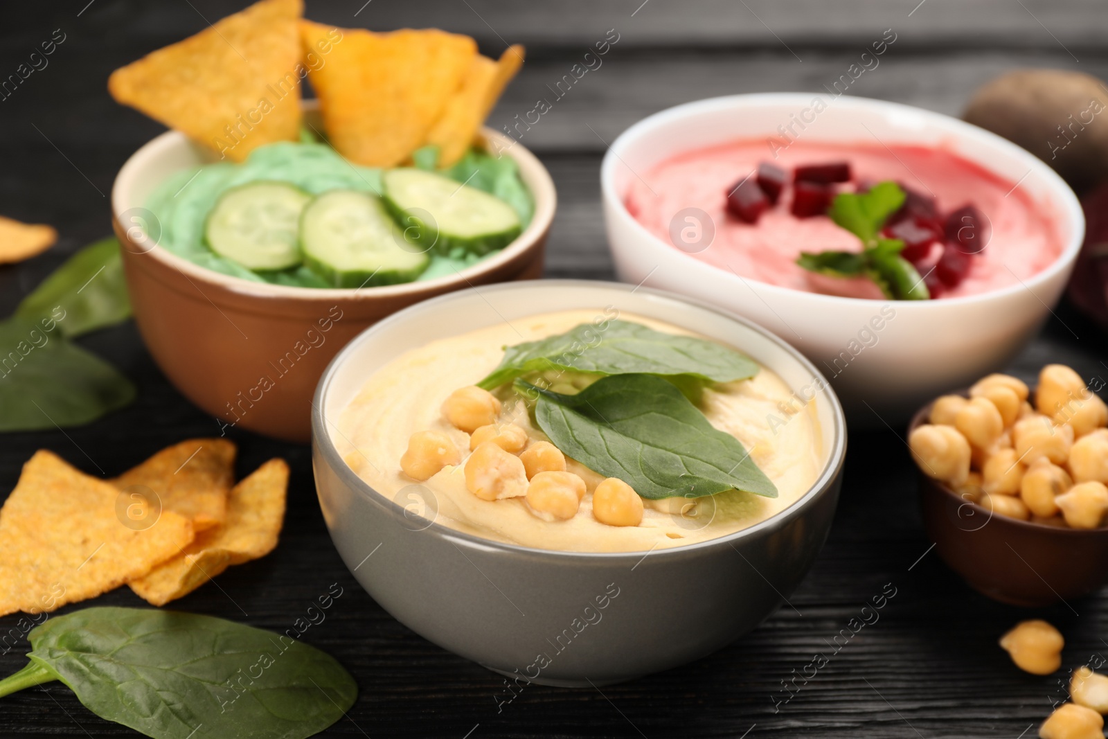 Photo of Different kinds of tasty hummus, nachos and ingredients on black wooden table