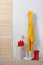 Photo of Red umbrella in holder, raincoat and rubber boots near white wall indoors