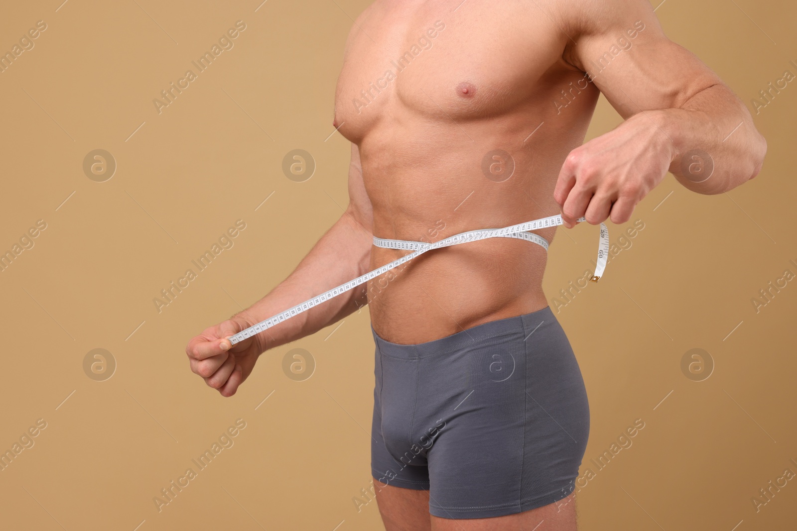 Photo of Athletic man measuring waist with tape on brown background, closeup. Weight loss concept