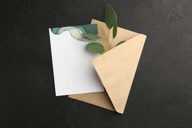 Photo of Envelope with blank invitation card and green leaves on black textured background, top view. Space for text