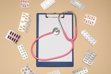 Photo of Stethoscope and clipboard surrounded by pills on beige background, flat lay. Medical tool