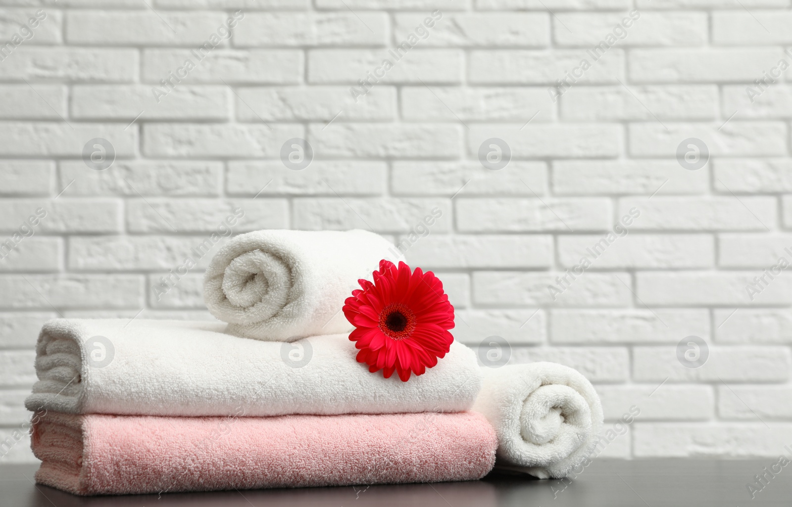 Photo of Rolled and folded soft towels with flower on table near brick wall. Space for text