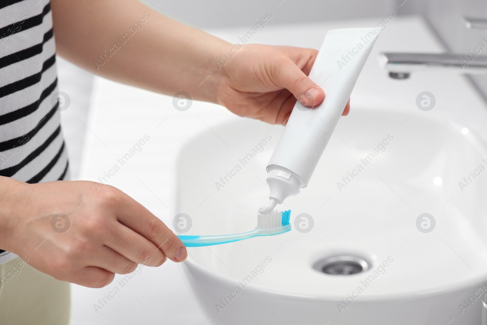 Photo of Woman squeezing toothpaste from tube onto toothbrush near sink in bathroom, closeup