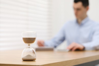 Hourglass with flowing sand on desk. Man using calculator indoors, selective focus