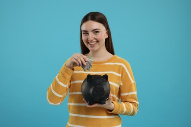 Happy woman putting dollar banknotes into piggy bank on light blue background