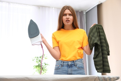 Emotional woman with iron and clothes at home