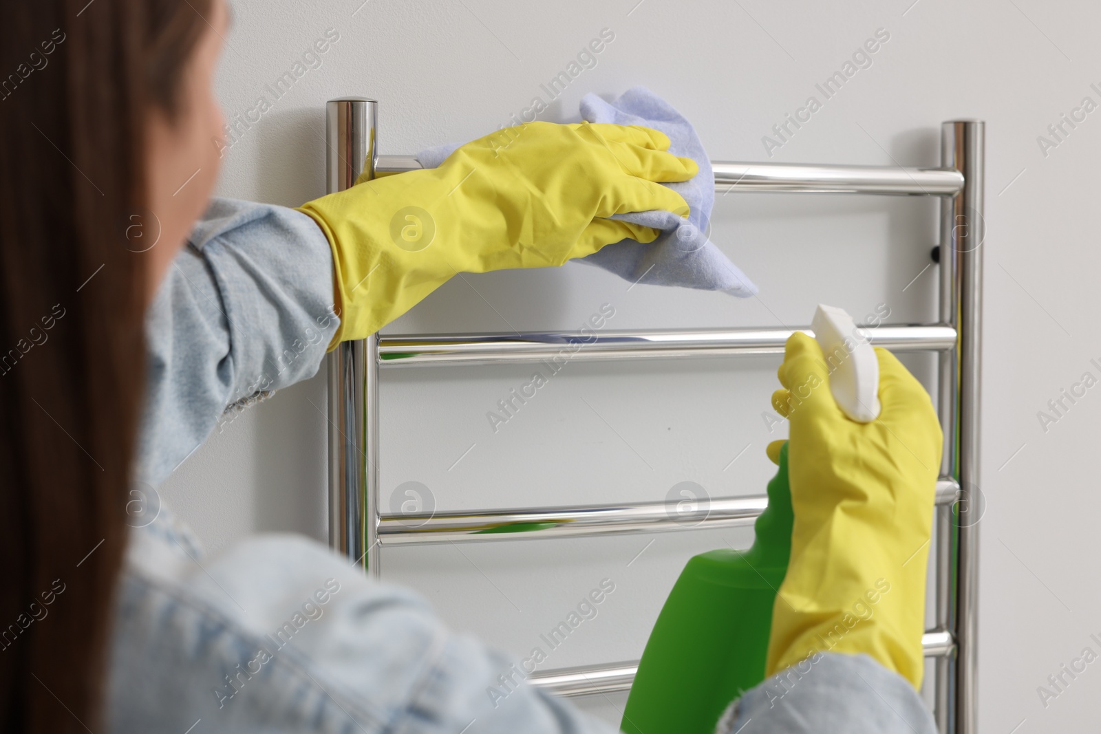 Photo of Woman cleaning heated towel rail with sprayer and rag, closeup