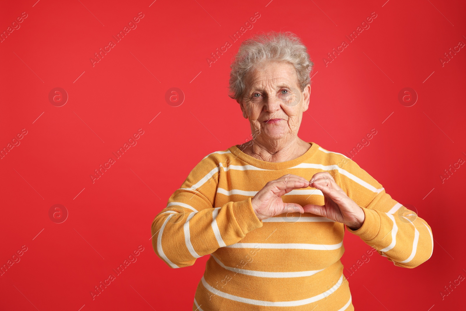 Photo of Elderly woman making heart with her hands on red background, space for text