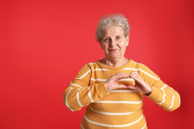 Elderly woman making heart with her hands on red background, space for text