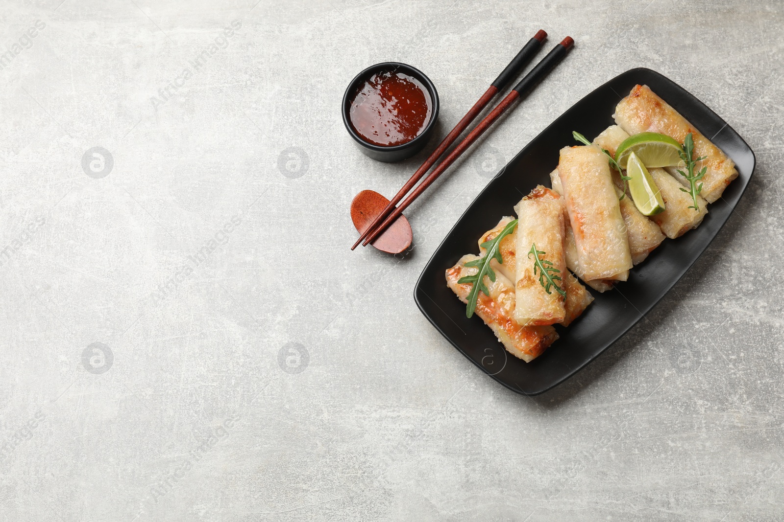 Photo of Tasty fried spring rolls, arugula, lime and sauce served on grey textured table, flat lay. Space for text