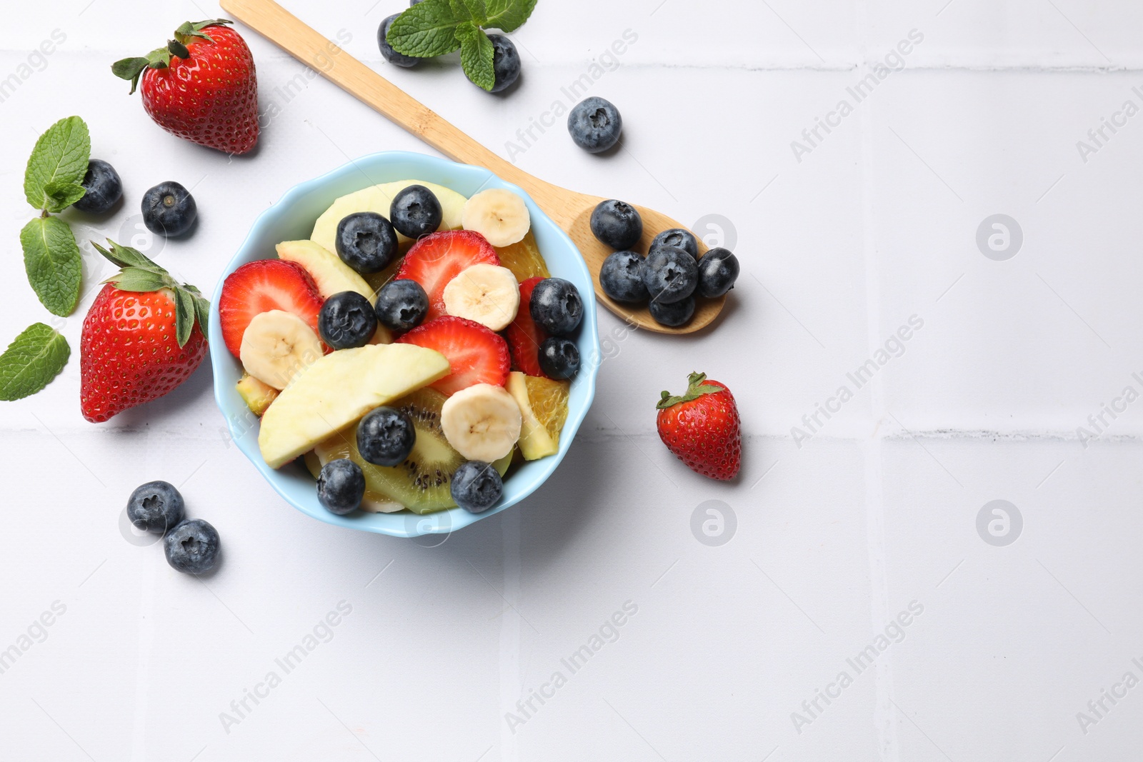 Photo of Tasty fruit salad in bowl and ingredients on white tiled table, flat lay. Space for text
