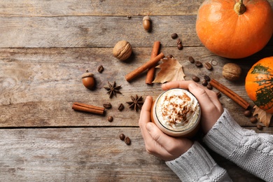 Woman holding glass of tasty pumpkin spice latte on wooden table, flat lay with space for text