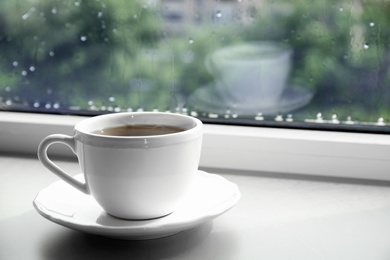 Photo of Cup of hot drink on window sill against glass with rain drops, space for text