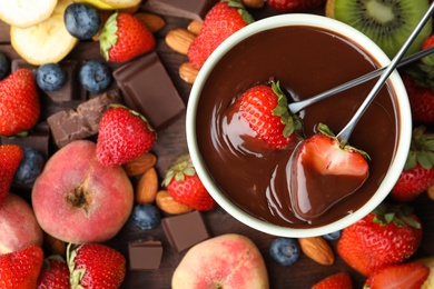 Photo of Fondue forks with strawberries in bowl of melted chocolate surrounded by other fruits on wooden table, flat lay