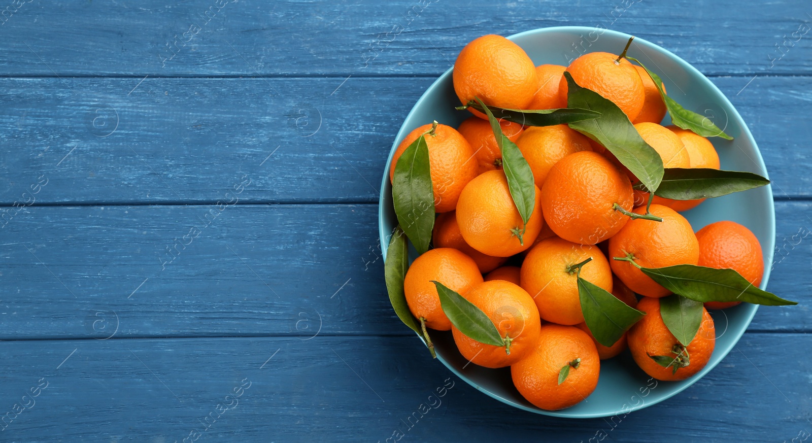 Photo of Fresh ripe tangerines with green leaves on blue wooden table, top view. Space for text
