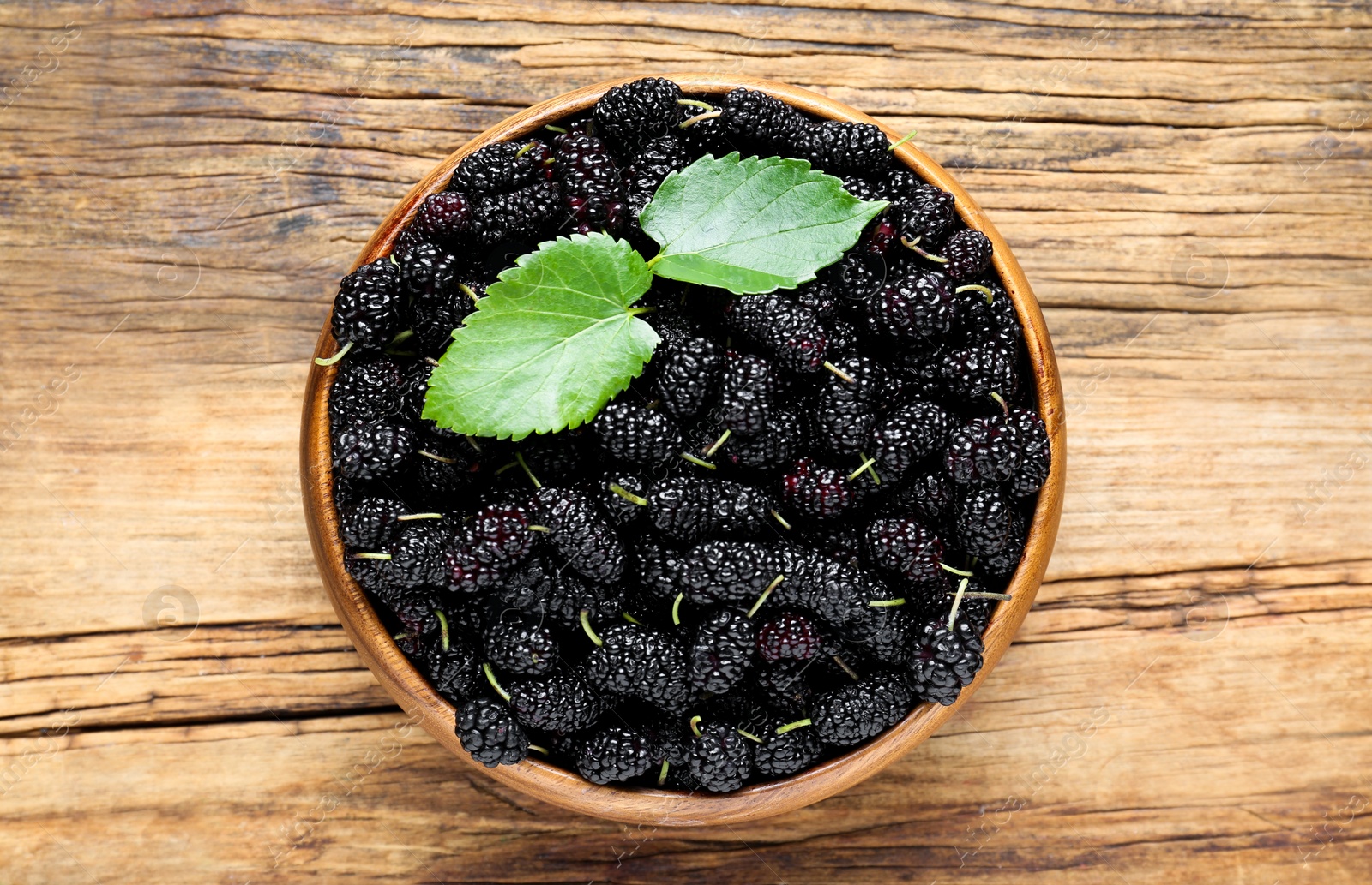 Photo of Fresh ripe black mulberries and leaves in bowl on wooden table, top view