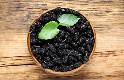 Fresh ripe black mulberries and leaves in bowl on wooden table, top view