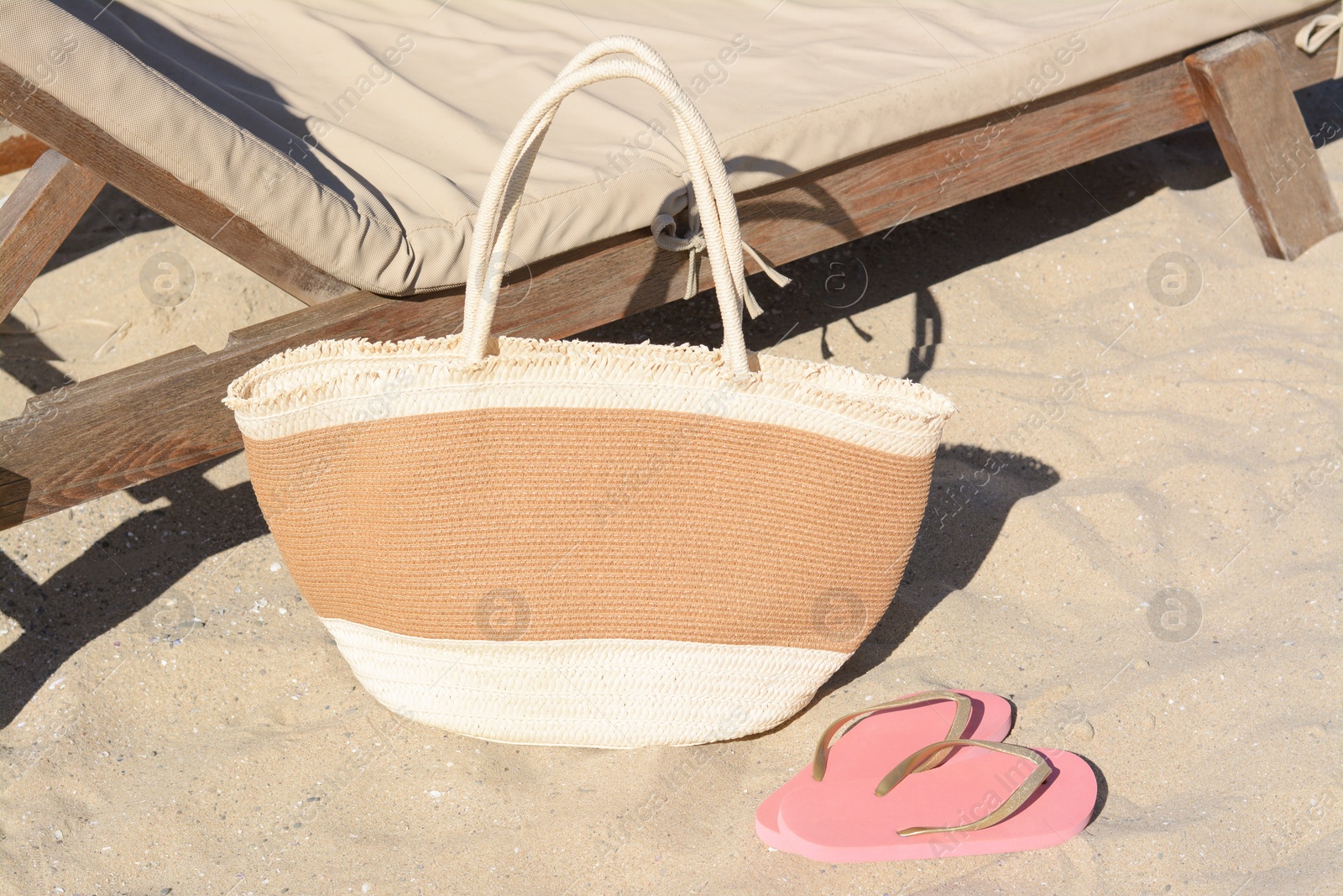 Photo of Straw bag and flip flops near wooden sunbed on sandy beach. Summer accessories